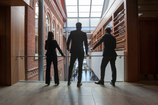 3 students standing on a balcony looking over the the bustling Smith foyer